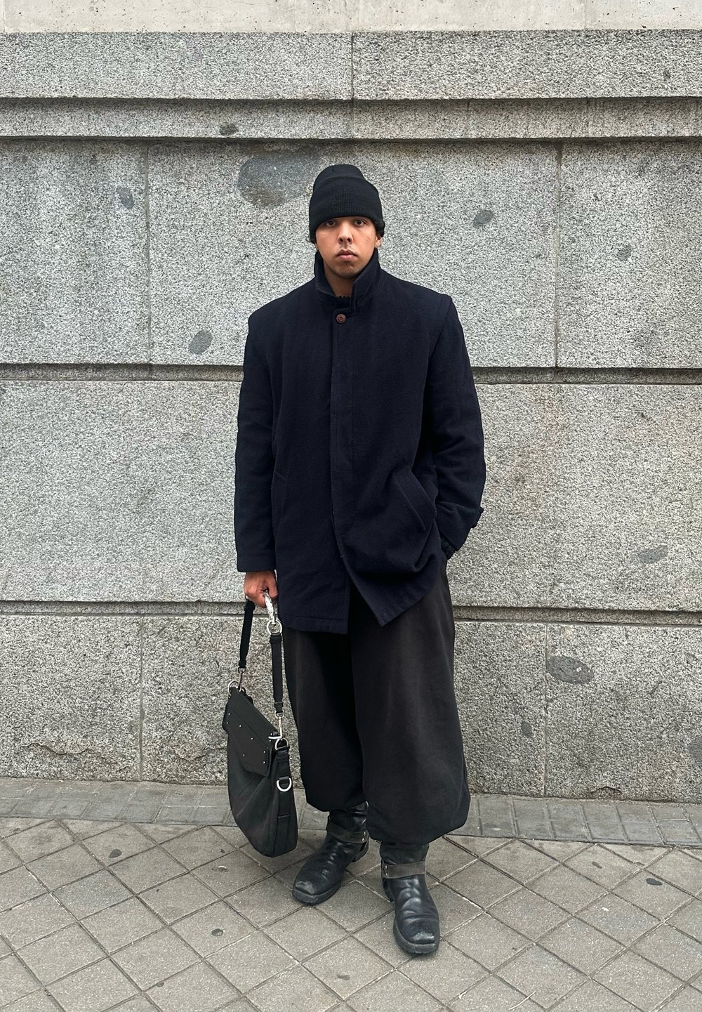 Μαύρο παλτό από μαλλί με ψηλό γιακά και κούμπωμα με κουμπιά, συνδυασμένο με φαρδιά μαύρα παντελόνια και μαύρες μποτάκια μέχρι τον αστράγαλο, κρατώντας μία μαύρη τσάντα ώμου.