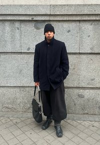 Μαύρο παλτό από μαλλί με ψηλό γιακά και κούμπωμα με κουμπιά, συνδυασμένο με φαρδιά μαύρα παντελόνια και μαύρες μποτάκια μέχρι τον αστράγαλο, κρατώντας μία μαύρη τσάντα ώμου.