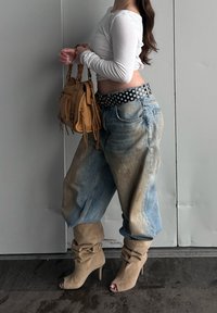 Jeans larges de couleur claire avec des zones délavées, ceinture à pois, haut blanc à manches longues, et bottes beige décontractées montant au-dessus du genou avec talons. Sac à main marron.