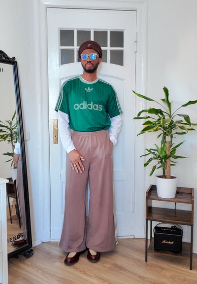 Jersey Adidas a rayas verdes y verticales con logo gris, camisa blanca de manga larga, pantalones anchos beige y zapatos planos burdeos. Entorno interior.