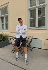 Camisa blanca de encaje con botones, pantalones cortos a rayas, zapatillas grises y calcetines blancos, sentado en una mesa de café cerca de una pared y una ventana.