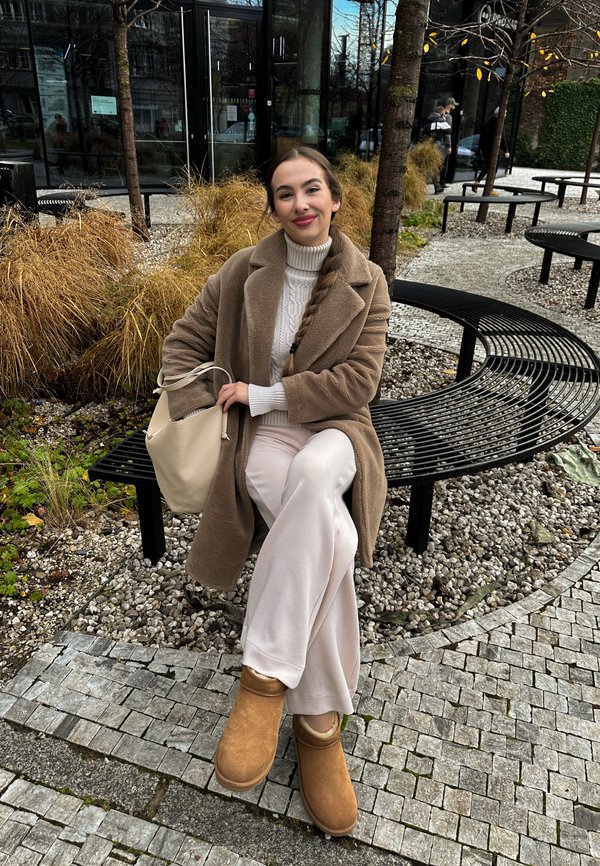 Beige faux fur coat, cable-knit cream sweater, light beige wide-leg pants, brown suede ankle boots, holding a cream handbag, seated on a black bench.