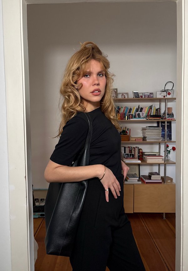 Jeune femme aux cheveux bouclés blonds portant une tenue noire et un grand sac à épaule noir, debout à l'intérieur près d'une étagère en bois remplie de livres et de décoration.