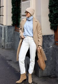 Manteau long beige, col roulé bleu clair, jean blanc et bottines beiges. Tient un sac marron texturé ; se tient contre un mur de pierre.