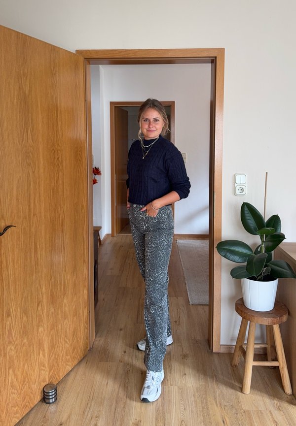 Giovane donna con pantaloni fantasia e maglione scuro in piedi in una porta, con una pianta in vaso su uno sgabello di legno vicino.