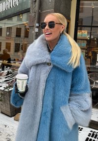 Ψεύτικο παλτό γούνας με χρώμα που μεταβάλλεται από ανοιχτό μπλε σε γκρι, διαθέτοντας έναν μεγάλο γιακά, κλείσιμο με έναν μόνο κουμπί και πλαϊνές τσέπες. Κρατάει μία κούπα καφέ.