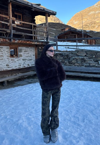 Mujer con chaqueta de piel oscura, pantalones de camuflaje y gafas de sol, de pie sobre la nieve cerca de casas rústicas de piedra y madera en una zona montañosa.