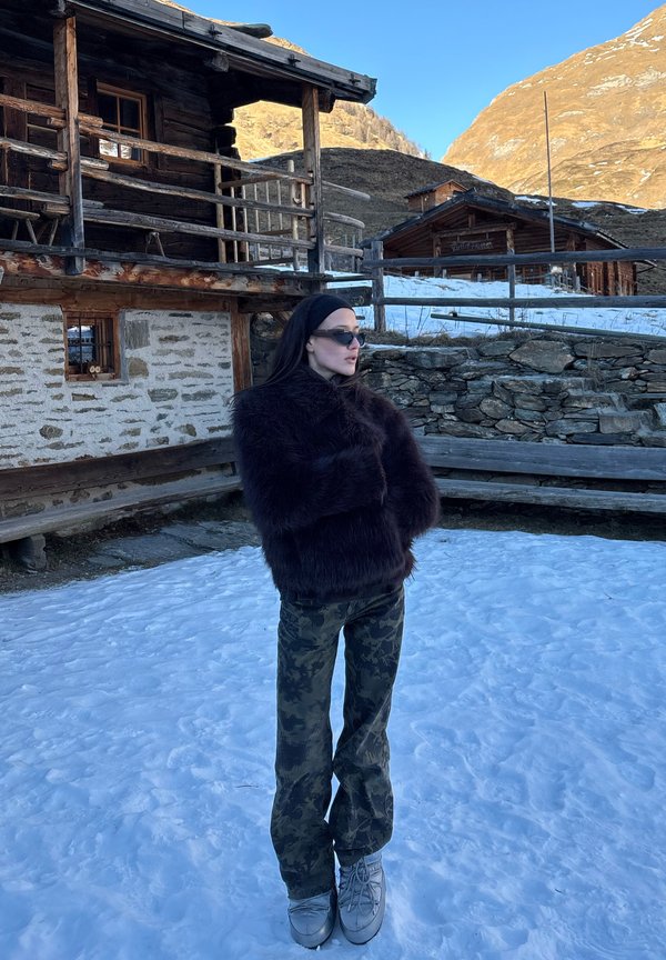 Γυναίκα με σκούρο γουνάκι, παντελόνι παραλλαγής και γυαλιά ηλίου στέκεται στο χιόνι κοντά σε παραδοσιακά πέτρινα και ξύλινα σπίτια σε ορεινή περιοχή.