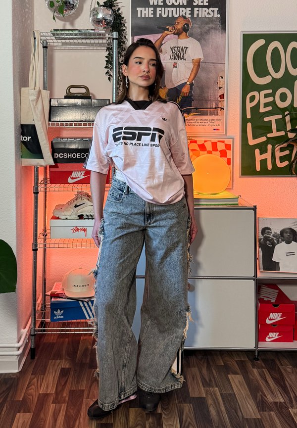 Młoda kobieta w koszulce ESPN i podartym dżinsie stoi w pomieszczeniu z półkami na sneakersy, oprawionymi plakatami i ciepłym oświetleniem.