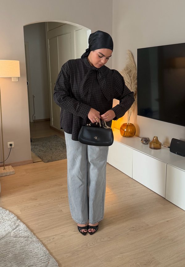 Mujer con pañuelo negro en la cabeza y blusa de cuadros sosteniendo un bolso negro de Calvin Klein en una sala de estar con suelo de madera y decoración moderna.