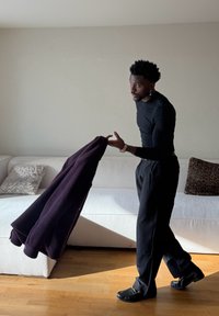 Homme en vêtements noirs tenant une veste violet foncé debout près d'un canapé blanc avec des coussins à motifs dans une pièce éclairée par le soleil.