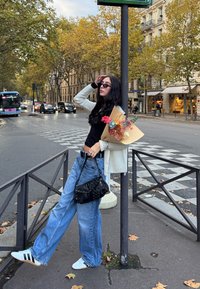 Vrouw met zonnebril leunt op een paal, houdt een bloemenboeket en een zwarte handtas vast, staand op een stadsstraat met herfstbomen en verkeer op de achtergrond.