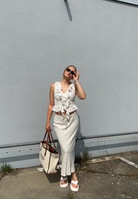 Weißes, blumengemustertes Crop-Top mit Rüschen, kombiniert mit einem langen, hellen Rock. Die Accessoires umfassen dunkle Sonnenbrillen und eine Einkaufstasche.