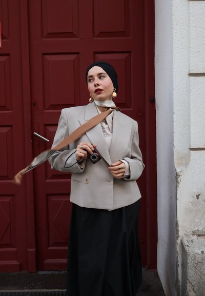 Blazer de gran tamaño en gris claro con hombros pronunciados, combinado con una blusa crema y una falda negra fluida. Pendientes en tono dorado y una cámara complementan el look.