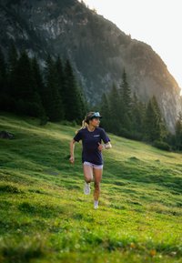 Idrettsutøver i en mørkeblå lettvekts skjorte og hvite shorts som løper på gress med fjell og furutrær i bakgrunnen; solfylt scene.