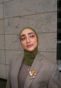 Olivengrønn hijab, lysegrå blazer, brun topp under, og en gullfarget blomsterbrosje som aksent. Jevn tekstur og skreddersydd passform.