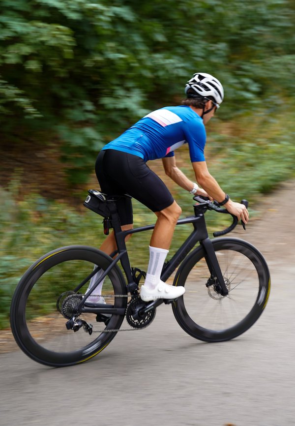 Cyclist wearing blue jersey, black shorts, and white helmet rides road bike on paved path with greenery in background.