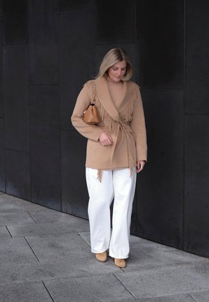 Chaqueta cruzada beige con detalles de flecos, combinada con pantalones blancos de pierna ancha y tacones beige. Sosteniendo un pequeño bolso marrón, de pie sobre un pavimento de piedra.