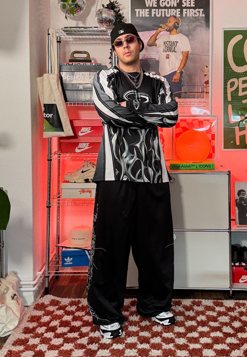 Homme portant une chemise noire à motifs de flammes blanches et un pantalon ample, debout les bras croisés dans une pièce avec des boîtes de baskets et des affiches.