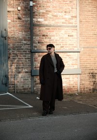 Homme portant un long manteau sombre, un pull gris, des gants noirs et des lunettes de soleil, debout sur le trottoir contre un mur de briques éclairé par le soleil.