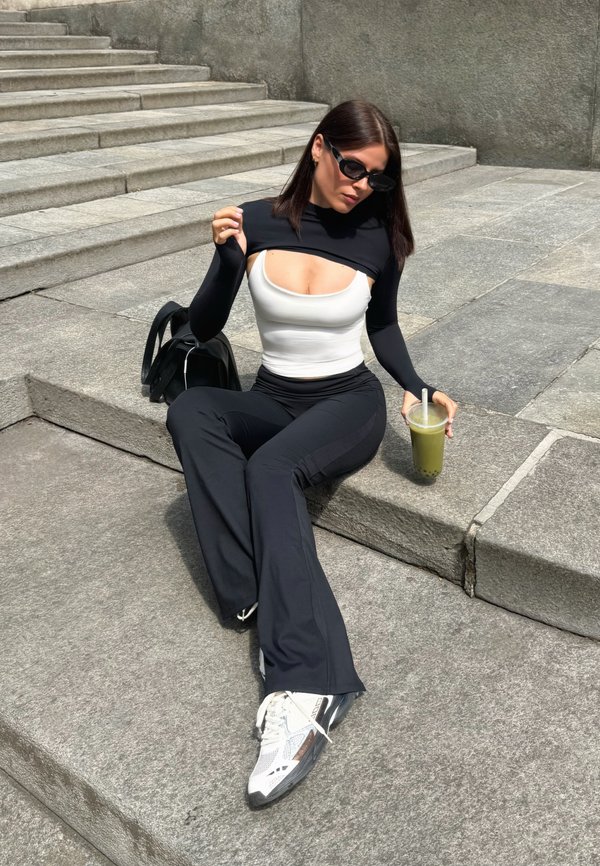 Jonge vrouw in een zwart-witte outfit zit op betonnen trappen, draagt een zonnebril en houdt een groen bubble tea drankje met een rietje vast.