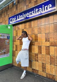 Mladý muž s copánky, oblečený v bílém tílku a šedých šortkách, stojí opřený o obloženou zeď pod nápisem Cité Universitaire.