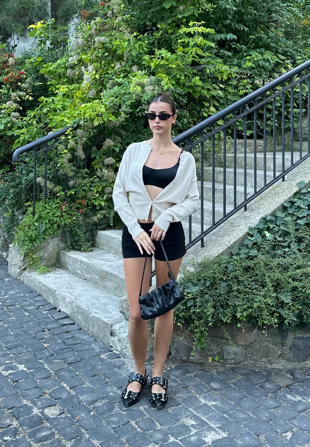 Λευκό πουκαμισάκι με κουμπιά, μαύρο κοντό τοπ και εφαρμοστό μαύρο φούστα. Μαύρα παπούτσια με αγκράφες. Μικρή μαύρη τσάντα. Πράσινη φυλλωσιά στο φόντο.