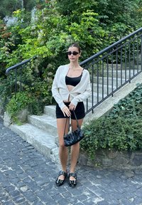 Λευκό πουκαμισάκι με κουμπιά, μαύρο κοντό τοπ και εφαρμοστό μαύρο φούστα. Μαύρα παπούτσια με αγκράφες. Μικρή μαύρη τσάντα. Πράσινη φυλλωσιά στο φόντο.