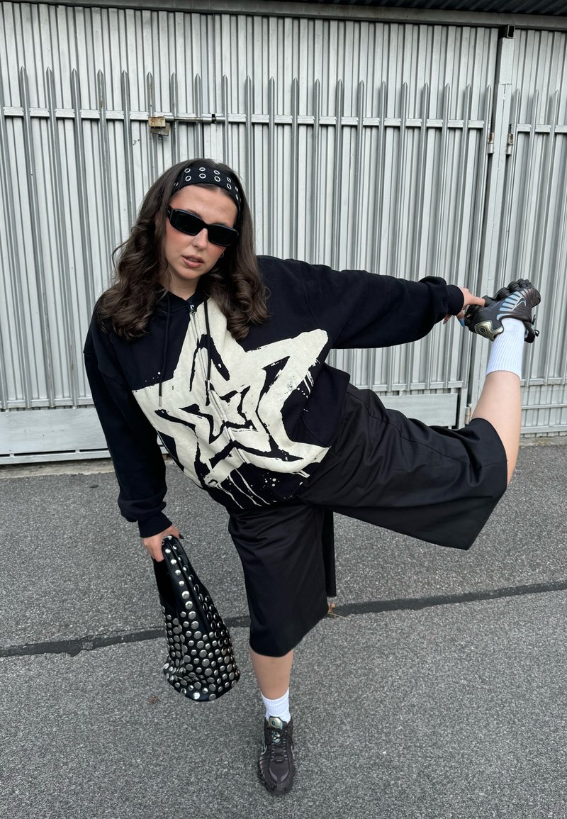 Sweatshirt preto com gráfico de estrela branca, calções pretos até ao joelho, saco preto texturizado com tachas, ténis escuros, meias brancas até ao tornozelo, óculos de sol pretos.