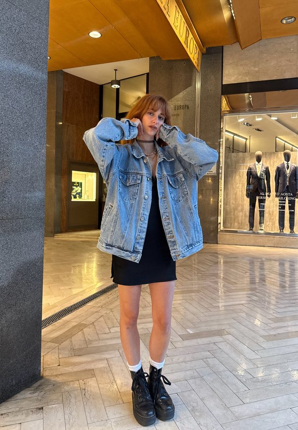 Mlada ženska z rdečimi lasmi, oblečena v prevelik jeans jakno, črno obleko, bele nogavice in črne platfrom čevlje, stoji v zaprtih prostorih blizu trgovine s suknjiči.