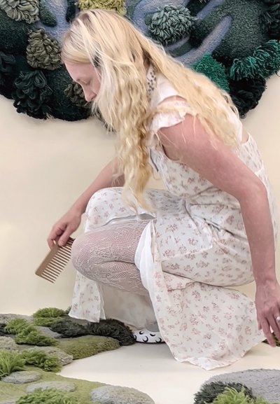 Vestido floral beige sobre capas blancas, medias texturizadas, sosteniendo un peine de madera, arrodillada cerca de formas de alfombra acolchada en verde y gris.