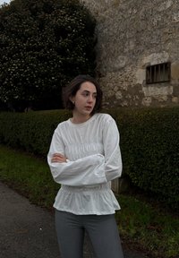 Λευκή μπλούζα με μακριά μανίκια και πτυχώσεις στη μέση, συνδυασμένη με στενές γκρι παντελόνες. Στο φόντο διακρίνεται μια φράχτη και ένας τοίχος από πέτρα.