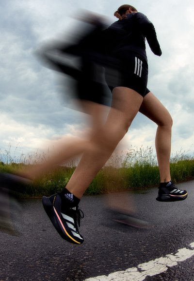 Zapatillas deportivas negras con rayas blancas, acentos en la entresuela y agarre texturizado, llevadas con pantalones cortos negros, en movimiento sobre una carretera.