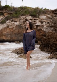 Acoperire din tricot, semi-transparentă, de culoare bleumarin, cu un design cu umeri decupați, model texturat și fante laterale, purtată pe plajă, cu apă stropind în jur.