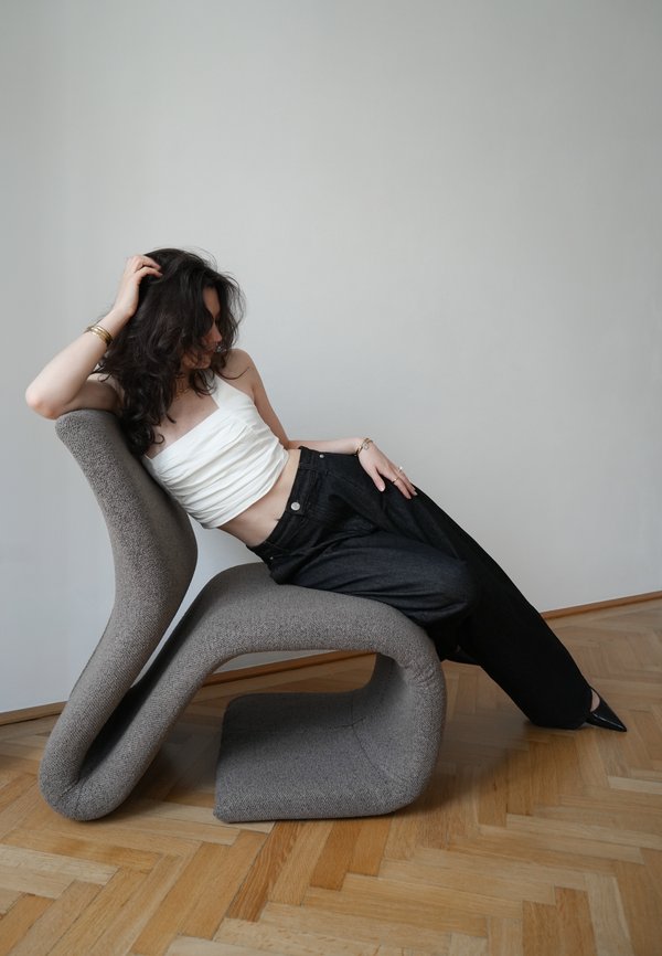 Femme aux cheveux foncés se détendant sur une chaise moderne grise incurvée, vêtue d'un haut blanc, d'un pantalon noir et de chaussures noires pointues sur un sol en bois.
