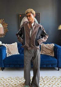 Henkilö, jolla on kiharat vaaleat hiukset ja aurinkolasit, ruskea ja musta takki sekä harmaat housut, seisoo olohuoneessa, jossa on sininen sohva ja kuvioidut tyynyt.
