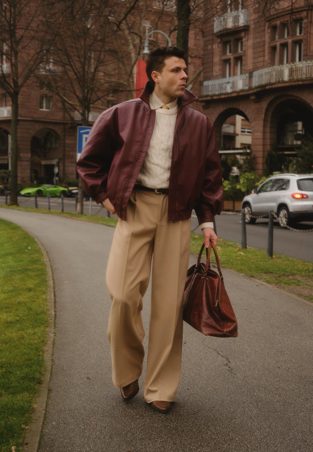 Veste aviateur en cuir marron rouge, pull en maille torsadée crème, pantalon large beige, chaussures marron et sac à main marron, dans un environnement urbain.