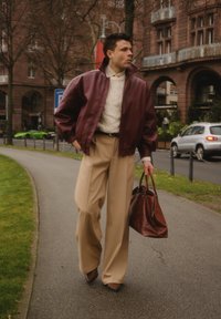Veste aviateur en cuir marron rouge, pull en maille torsadée crème, pantalon large beige, chaussures marron et sac à main marron, dans un environnement urbain.