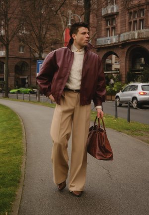 Veste aviateur en cuir marron rouge, pull en maille torsadée crème, pantalon large beige, chaussures marron et sac à main marron, dans un environnement urbain.
