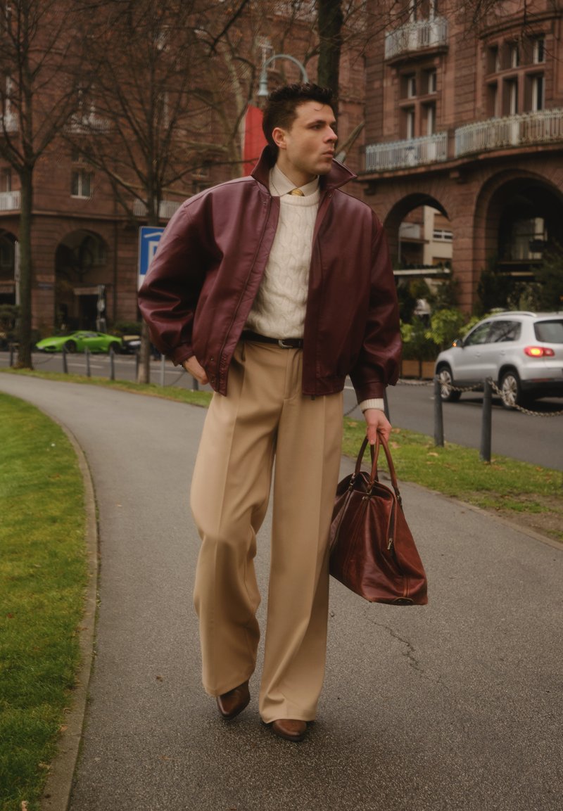 Giacca bomber in pelle color marrone scuro, maglione a trecce color crema, pantaloni beige a gambe larghe, scarpe marroni e borsa marrone, ambientati in un contesto urbano.