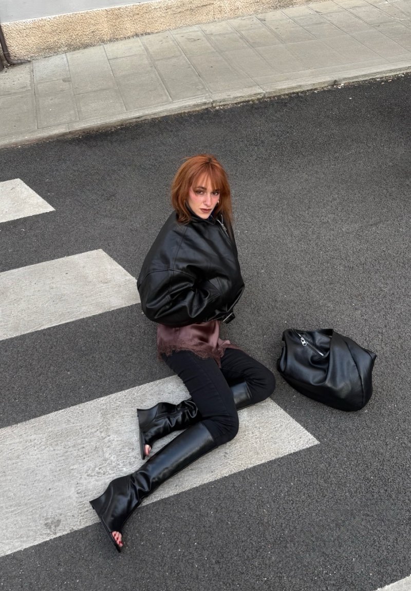 Frau mit roten Haaren, die eine übergroße schwarze Jacke und hohe Stiefel trägt, kniet auf einem Zebrastreifen, eine schwarze Tasche liegt neben ihr.