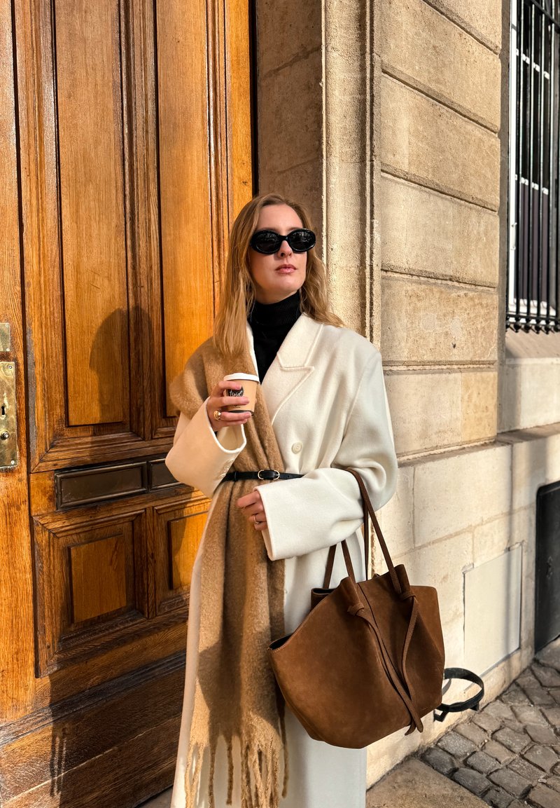 Manteau en laine blanche porté par-dessus un col roulé noir, avec une écharpe beige drapée. Tenant une tasse de café et portant un sac en daim marron. Fond de porte en bois.