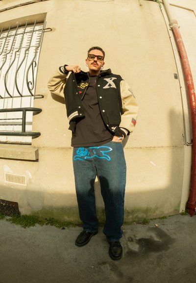 Chaqueta universitario negra y beige con gráfico, camiseta negra, jeans azules con grafitis que presentan un diseño de caricatura azul, calzado negro.