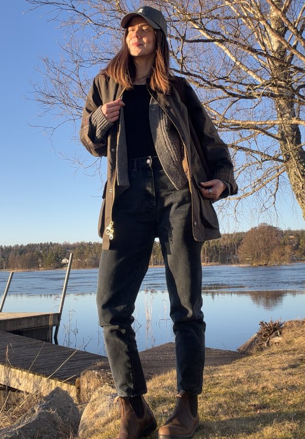 Kvinde iført mørk jakke, jeans, støvler og kasket står ved en sø med bare træer og en træbro i sollys.