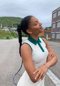 Robe sans manches blanche à côtes avec un col vert foncé et des boutons. Grande tresse noire et bracelet en argent. Fond gris avec de la verdure.