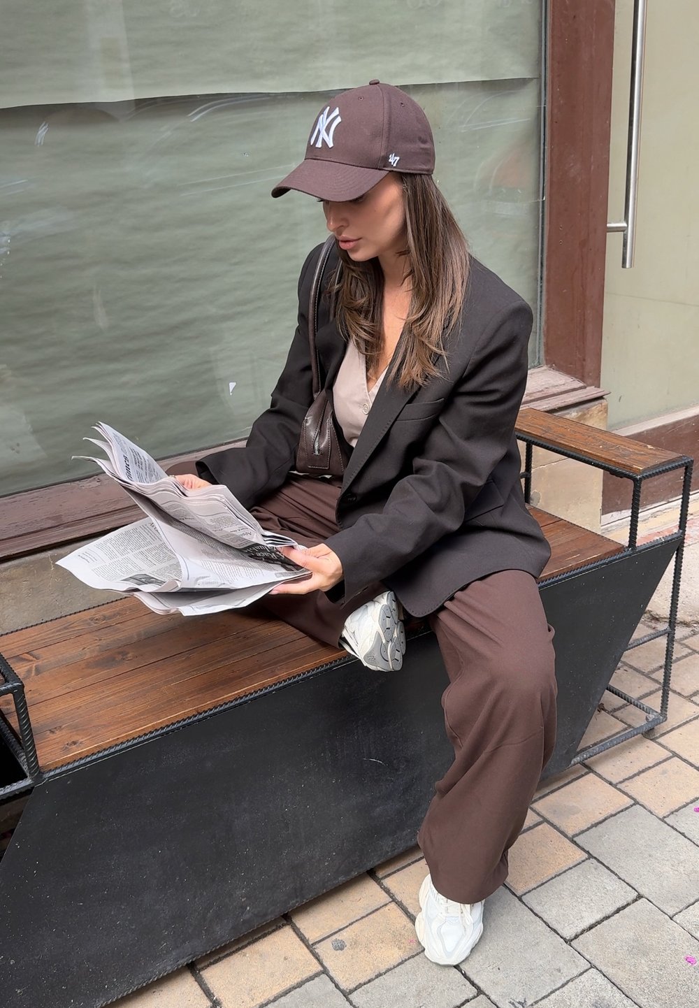 En person i en sort oversized blazer sidder på en træbænk og læser aviser. Personen har en brun kasket på, en beige top og brune bukser med hvide sneakers.