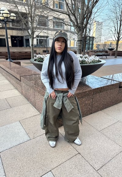 Pantalones cargo de color verde oliva con corte oversize, suéter de canalé gris y gorra de camuflaje, de pie al aire libre cerca de un arreglo floral.