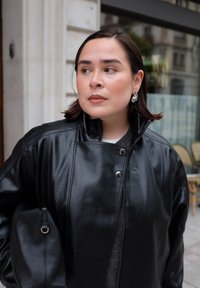 Młoda kobieta w czarnej skórzanej kurtce i srebrnych kolczykach, trzymająca czarną torebkę, patrząca w bok na ulicy miejskiej.