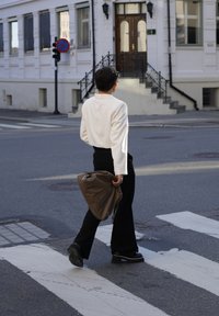 Ένα άτομο με κρεμ σακάκι και μαύρο παντελόνι περπατά σε διάβαση πεζών, κρατώντας μια καφέ τσάντα με ματ υφή. Στο φόντο διακρίνεται ένας δρόμος και ένα κτήριο.