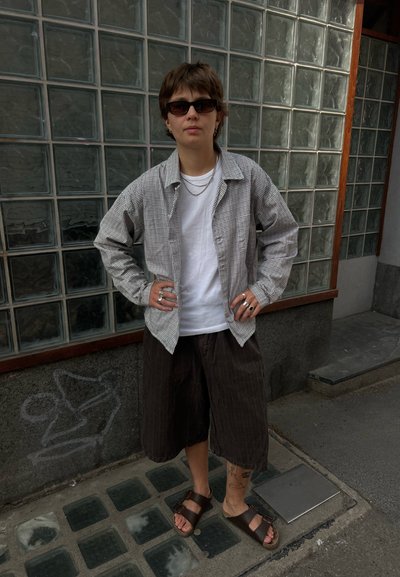 Persona con gafas de sol, chaqueta de cuadros, camisa blanca, pantalones cortos oscuros y anchos y sandalias, de pie con las manos en las caderas contra una pared de bloques de vidrio.